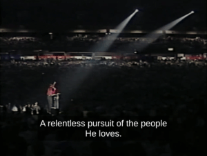 Ron Luce stands at a podium in front of tens of thousands of teens. The caption reads, "A relentless pursuit of the people He loves." A continuation of his speech about how we're pursuing Jesus relentlessly. He shakes his fist to punctuate each statement while the crowd grows wild.