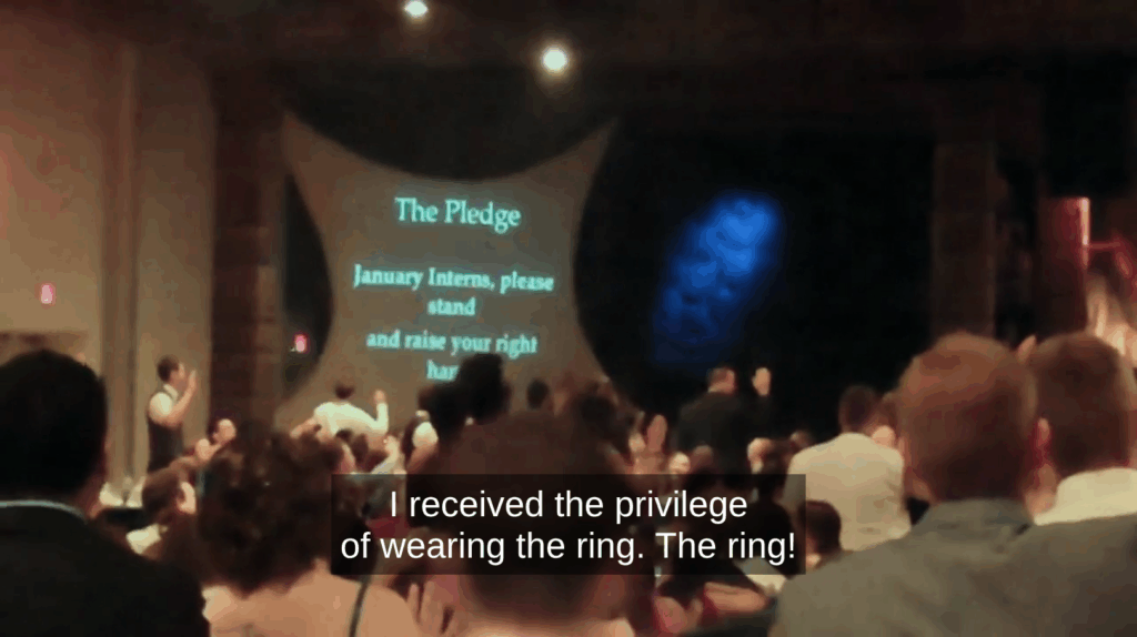 A gathering of January (2001?) interns in the Teen Mania auditorium getting ready to take the Pledge where they don their honor rings. The interns are holding their right hands up, as commanded by the screen. The caption reads, "I received the privilege of wearing the ring. The ring!" where the interns shout "The ring!" as part of the chant: "The ring! The line! Always honorable, for life!" The caption cuts off before the rest of the chant.