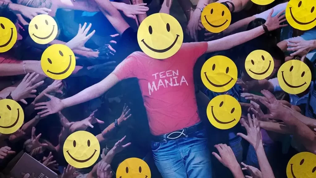 An individual clad in jeans and a tucked-in, red t-shirt emblazoned with "Teen Mania" crowd surfs. Everyone's face is blocked with Shiny Happy People's iconic yellow smiley faces.